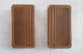 Zwei große rechteckige braune Boxen, die vor einer weißen Backstein-Wand an weißen Fäden hängen. Unten hängen weiße Kabel raus. Um die Holzkörper führen goldene Rohrstangen, an denen die Boxen aufgehängt sind. 