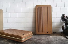 Zwei Musik-Boxen, eine lehnt an einer weißen Mauerwand, eine liegt auf dem Boden. Die braunen Boxen haben längliche Schlitze, mehrere Holzschichten und goldene Rohrstangen, die einmal ganz herum führen. 