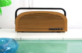 Ein braunes, längliches Radio am Rand eines Pools mit türkisem Wasser. Das Radio ist mit goldenen Schrauben zusammengeschraubt, hat längliche, horizontale Schlitze und einen schwarzen Röhren-Griff, der über die gesamte Radio-Länge geht. Unten rechts steht: Sunset 1, mit schwarzen Schreibmaschinen-Tasten. Das Radio steht auf weißen Platten. Links im Pool ein Stück von einer aufblasbaren Palme. 