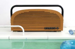 Braunes Radio mit länglichem Rohr-Griff und länglichen Schlitzen. Der Name Sunset 1 steht rechts unten in Schreibmaschinen-Tasten. Weiße Bodenplatten, davor türkises Wasser, daneben eine blaue Taucherbrille und ein blauer kleiner Schwimmring. 