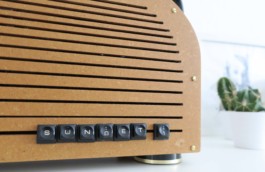 Detail-Ansicht der selbstgebauten Boombox Sunset 1. Der Name steht mit schwarzen Schreibmaschinen-Tasten rechts unten. Darunter ein schwarz-goldener Standfuß. Goldene Schrauben. Im Hintergrund ein Kaktus und eine Porträt-Zeichnung. 