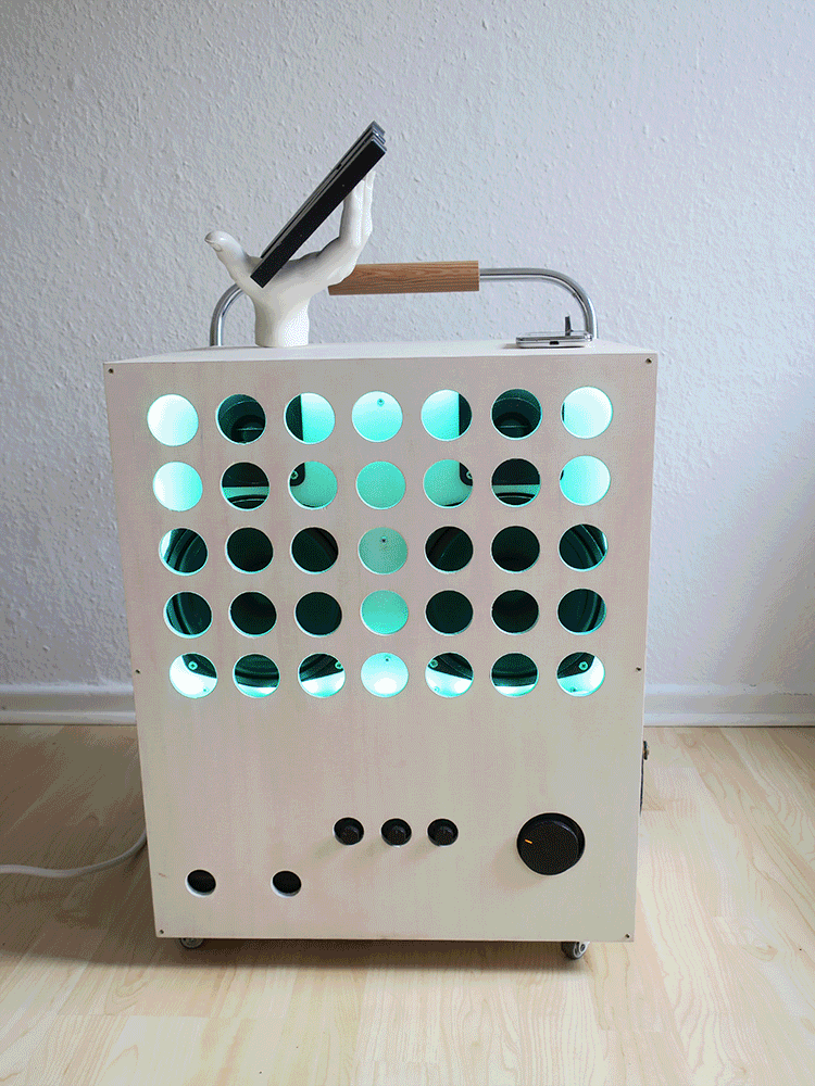 Selbstgebaute Boombox für den Handball-Verein SV Wersten 04. Eine weiße Box auf Rädern, mit schwarzen Knöpfen und runden Löchern, hinter denen 2 große schwarze Boxen zu sehen sind und das Licht die Farbe wechselt. Oben ein Tragegriff mit Holz. Und eine Schaufensterhand mit einem schwarzen Holz-W, die sich dreht. 