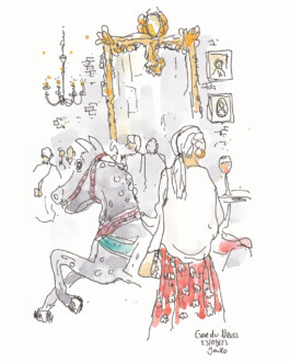 Eine Frau steht an einem Stehtisch, auf dem ein Glas Wein steh. Der Stehtisch ist aus einem alten Karussell-Pferd gemacht. Im Hintergrund ist eine alte Backsteinwand mit alten Bildern und einem goldenen Spiegel zu sehen.